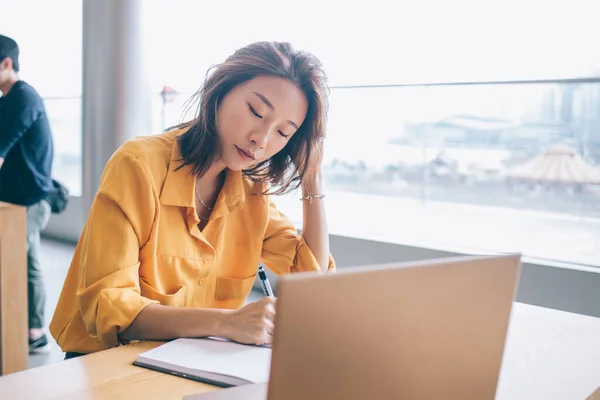 Asian female designer in yellow blouse drawing in notebook and touching hair at desk with laptop at workplace with panoramic windows in Hong Kong city