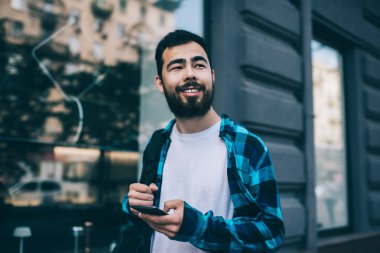 Sıradan giyinmiş neşeli Asyalı hippi sokakta 4G internete bağlı cep telefonunu tutuyor. Yolculuk sırasında navigasyon için akıllı telefon uygulaması kullanan mutlu milenyum erkek yolcusu.