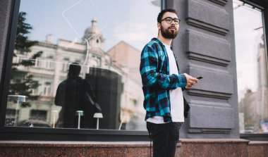 Bin yıllık yakışıklı Asyalı sıradan giyinen, arka planda duran cep telefonlu genç sakallı hippi, şehirdeki sosyal ağlarda sohbet etmek için akıllı telefon kullanıyor.