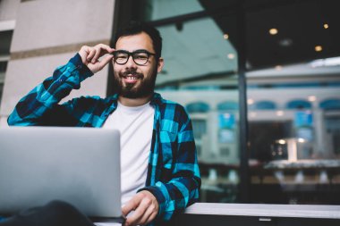 Görüş düzeltmesi için göz makyajı yapan neşeli sakallı serbest erkek portresi dışarıda oturmaktan memnun olan mutlu Asyalı adam dijital göçebe dizüstü bilgisayar ve 4G internet kullanıyor.
