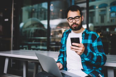 Gözlüklü Asyalı erkek, akıllı telefondan para transferi bildirimini kontrol ediyor. İnternette bilgisayarla uzaktan çalışan bir iş var. Milenyum hipster 'ı cihazları senkronize ediyor.