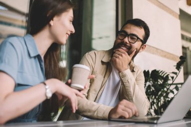 Kafeteryada dizüstü bilgisayarın yanında oturan beyaz kadın arkadaşların konuşmalarını dinleyen Asyalı sakallı mallara odaklan, pozitif genç meslektaşlar Brea hakkında sohbet ediyorlar.