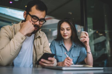 Asyalı erkek öğrenci bilgi aramak için cep telefonunu kullanırken, Kafkasyalı kadın arkadaşıyla ödev hazırlarken, fotokopi defterine not alırken, çift kafeteryada oturup akıllı telefondan video izliyor. 