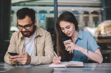 Molada kafeteryada birlikte oturan neşeli çok ırklı öğrenciler, not defterine beyaz kadın yazarken, Asyalı erkek akıllı fonla sosyal ağlardaki içeriği kontrol ediyor.