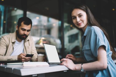 Laptop bilgisayarında çalışırken kameraya bakan çekici kadın ressamın portresi, Asyalı erkek meslektaşın yanında oturan, serbest çalışan güzel beyaz kadın.