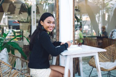 Bali 'deki kafenin terasında beyaz bir masada oturmuş kameraya bakarken akıllı telefonlu güzel Asyalı bir kadının yan görüntüsü.