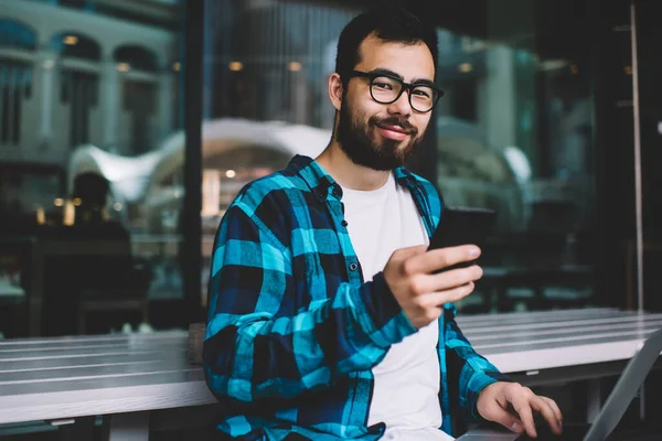 Portrait of successful asian male freelancer using laptop computer and smartphone during remote job, prosperous millennial hipster guy checking notification on mobile phone looking at camera outdoor