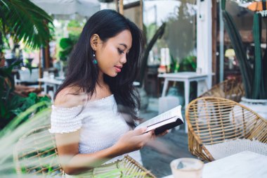 Beyaz omuzlu, kitap okuyan, açık terasta bir fincan kahveyle oturan Asyalı çekici bir kadının yan görüntüsü.