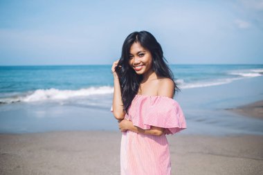Pembe elbiseli Endonezyalı genç bir kadın okyanusa ve gökyüzüne bakarak gülümsüyor. Bali sahilinde kameraya bakıyor ve saçlarına dokunuyor.