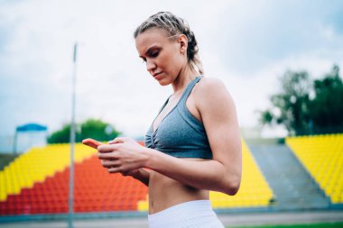Stadyumda elinde turuncu akıllı telefonuyla duran ve koşuya ara verilirken sosyal medyada gezinen konsantre ve odaklanmış bir bayan.