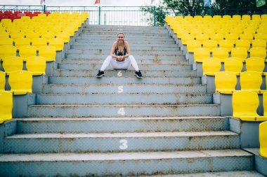 Spor kıyafetleri içinde keyifli bir kadın. Dostça gülümsüyor ve stadyumdaki eski püskü basamaklarda rahatlıyor ve kameraya bakıyor.