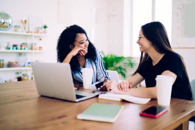 Gülümseyen uzun saçlı kadının geniş gülüşlü Afro-Amerikan bir kadınla konuşmasının yan görüntüsü. Dikkatlice dinleyen, elleri masada dizüstü bilgisayarlı ve akıllı telefonlu bir şekilde oturan bir kadın. 