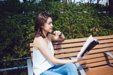 Kitap okurken çitlere yaslanmış kısa kahverengi saçlı, konsantre Asyalı genç kadının yan görüntüsü.