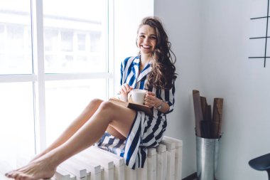 Çekici, çizgili elbisesiyle, elinde bir bardak sıcak içecek ve kitapla pencere kenarında çıplak ayakla kameraya bakarken çekici bir esmer.