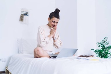 Beyaz ev kazağı giyen, bacak bacak üstüne atmış, el ele tutuşup, yatak odasında umut dolu dizüstü bilgisayara bakan bir kadın. 