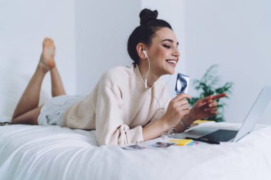 Yatakta dinlenen, elinde bilgisayarını gösteren ve evinde gülümseyen bir fotoğraf tutan mutlu genç kadının yan görüntüsü.