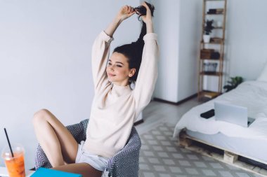 Düşünceli, güler yüzlü, beyaz tişörtlü, süveterli, modern koltukta dinlenen, vücudunun altında tik-tak eden bacağı olan, başını çeviren ve rahat yatak odasında bağlarını çözen saçları olan bir kadın.