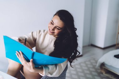 Aydınlık odada oturan kadın siyah beyaz sandalyede bej bir kazakla oturuyor ve mavi klasörde kağıda bir şeyler yazıyor.