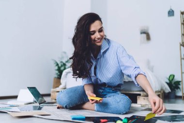 Uzun kıvırcık saçlı yaratıcı öğrenci. Siyah resimlere etiket yapıştırıyor. Dizüstü bilgisayarı ve bacak bacak üstüne atıyor.