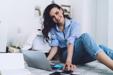 Modern odada akıllı telefonuyla oturmuş kameraya bakarken sıradan kıyafetlerle gezen mutlu bir kadın.