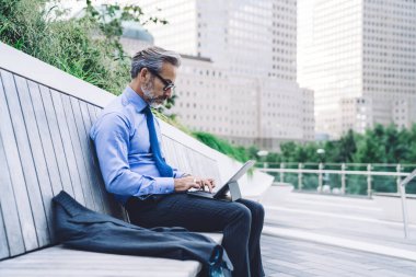 Orta yaşlı gözlüklü bir adamın tablet kullanırken tahta bankta New York caddesine karşı takım elbisesiyle oturuşunun yan görüntüsü.