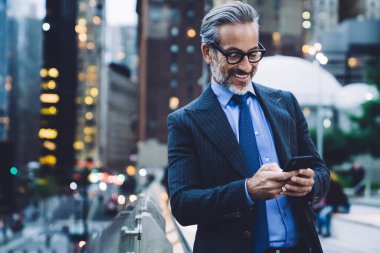 Yakışıklı, gri saçlı, mutlu iş adamı. Siyah takım elbiseli ve gözlüklü. New York 'un bulanık arka planında cam çitlerle geziyor.