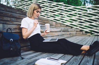 Resmi giysiler içinde, gözlüklü ve paket içkili yetişkin bir kadının ahşap merdivenlerde otururken dizüstü bilgisayar ekranına odaklanmış görüntüsü.