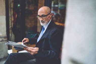 Gözlüklü, takım elbiseli, sakallı ve kahve fincanı olan, pencerelere gazete okuyan yaşlı, şık bir adamın yan görüntüsü.