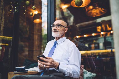 Akıllı telefonlu, kısa saçlı, gözlüklü, beyaz gömlekli, cadde kafesinin penceresine bakan yaşlı bir adam.