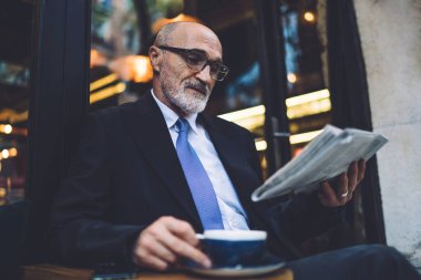 Kısa saçlı, sakallı, kahve bardağında gözlüklü, takım elbiseli, caddede oturan ve pencerelere gazete okuyan yaşlı, şık bir adam.