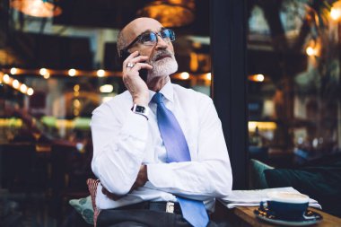 Gözlüklü, takım elbiseli, beyaz gömlekli, akıllı telefondan konuşan ve camdan dışarı bakan yaşlı yakışıklı bir adam.