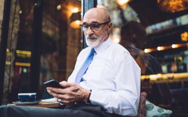 Gözlüklü ve beyaz gömlekli yaşlı kısa saçlı bir adam sokak kafesinin penceresinde oturuyor ve akıllı telefondan mesajlaşıyor.