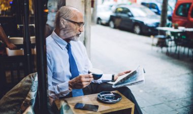 Beyaz gömlekli, yaşlı, kısa saçlı, kahve fincanı olan, yol kenarında oturan ve gazete okuyan bir adamın penceresinden.