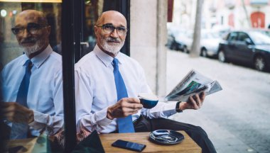 Beyaz gömlekli, elinde kahve ve gazete fincanı olan yaşlı, kısa saçlı bir adam yol kenarında oturuyor ve kameraya bakıyor.