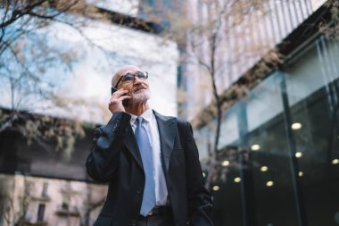 Kendinden emin, gri saçlı, resmi giyinmiş, gözlüklü bir adam cep telefonundan arıyor ve bina dışına bakıyor.