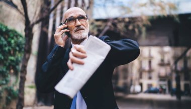 Yaşlı, kendine güvenen, beyaz saçlı, şık giyinen, elinde gazete olan, cep telefonuyla konuşan ve bina dışına bakan bir erkek.