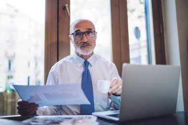 Gözlüklü, beyaz tişörtlü, elinde kahve fincanı ve kağıt çarşafla büroda kameraya bakarken.