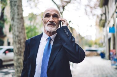 Resmi mavi takım elbiseli yaşlı, saygın girişimci. Düşünceli ve ciddi bir yüzü olan. Sokakta yürürken telefonla konuşan ve yüzünü çeviren.