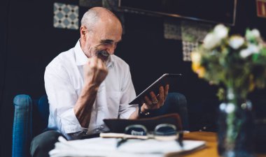 Beyaz tişörtlü, beyaz saçlı, neşeli kel yaşlı bir adam oturma odasında otururken yumruğunu kaldırmış tablet ekranına bakıyor.