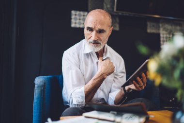 Kafası karışmış, kel, resmi giyinmiş, modern tableti karıştıran, kafenin masasındaki koltukta oturan, kafasını çevirmiş bir erkek.