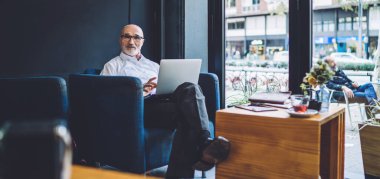 Kafede dizüstü bilgisayarın önünde oturan ve kameraya bakarken bacak bacak üstüne atan, resmi kıyafetli, kendinden emin bir iş adamı.