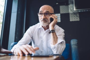 Kel, gri sakallı, gözlüklü, beyaz gömlekli, mavi sandalyede oturan ve telefonla konuşurken yüzünü çeviren yaşlı bir erkek.