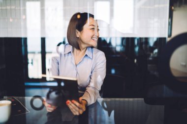 Klasik tişörtlü etnik bir kadın kahve molası veriyor gülümsüyor, dijital tablet kullanıyor ve pencereden dışarı bakıyor. 