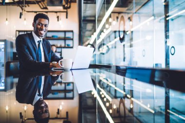 Klasik kıyafetli, beyaz dizüstü bilgisayarı ve bir fincan kahvesi olan şık siyah bir adam modern ofiste ışıklarla pencerenin yanında kırılıyor. 