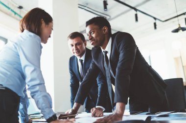 Çok ırklı genç meslektaşlar ellerinde cihazlar ve belgelerle masanın yanında duruyor ve şirket ofisindeki toplantı odasında proje planını tartışıyorlar.