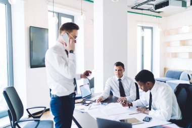 Gözlüklü ve şık elbiseli dalgın işadamının, gündüz vakti hafif modern ofisteki çok ırklı iş arkadaşları projesinde çalışmaya karşı kağıtlara yoğunlaşırken telefon görüşmesi yapması.