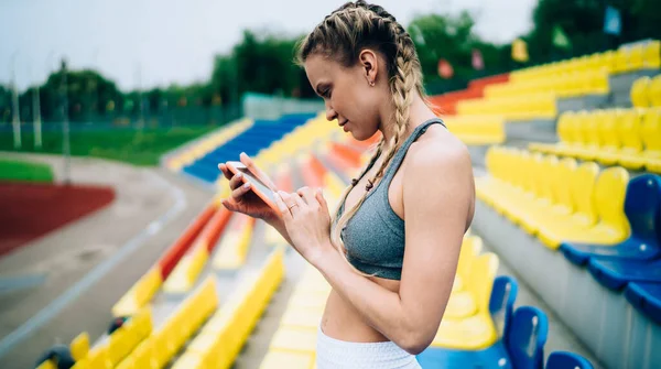 スポーツウェア姿のブロンドスポーツウェア姿の女性が スマートフォンでピグテールメッセージを発信しながら スタジアムのグランドスタンドに立っている — ストック写真