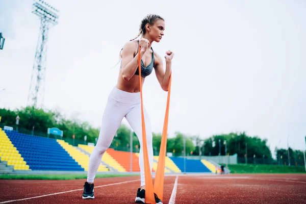 From below serious muscular female in sports clothes with pigtails training with rubber band at race track of open air stadium