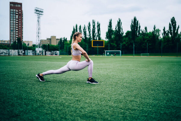 Side view of focused slender woman in modern sports clothing and comfy sneakers stretching by lunges at football field in city