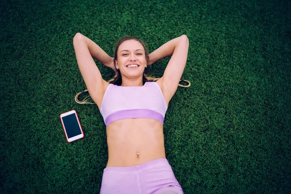 上から笑顔の幸せなスリム女性で快適な光スポーツウェアスマートフォンで芝生のフィールドに横たわっていると頭の後ろに手でカメラを見て — ストック写真